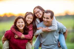 Family of four needing life insurance spend some time close together while outside. The Mother and Father each have an elementary school aged daughter on their back and they are smiling and enjoying a walk in the fresh air.